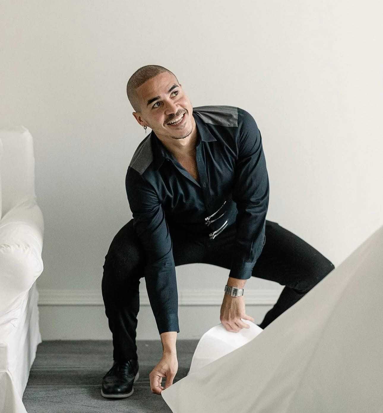 Man in black outfit lifting a sheet with a bright smile, in a minimalistic room setting.